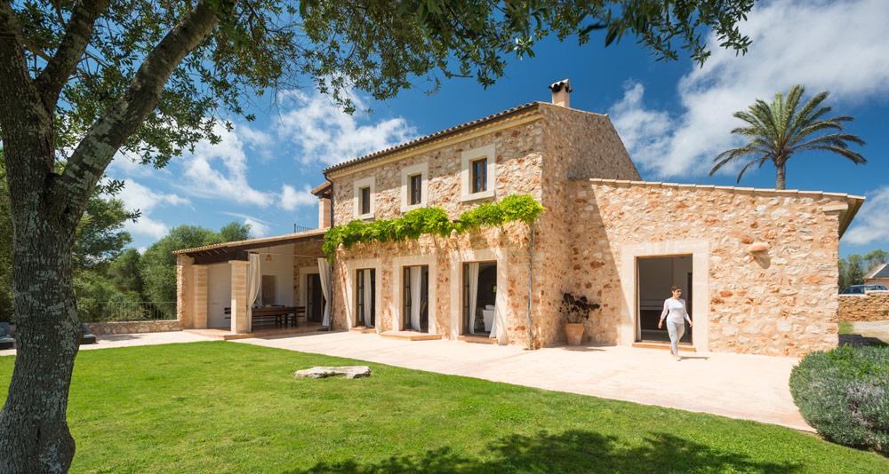 Ca'n Rossinyol, a two storey Mallorcan stone villa with a vine-covered facade, a covered terrace and a wide lawn under a blue sky